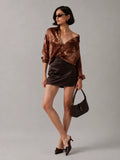 A model wearing a brown satin off-the-shoulder top, a dark brown satin mini skirt, and holding a dark brown handbag against a solid gray studio background.