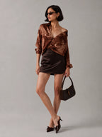 A model wearing a brown satin off-the-shoulder top, a dark brown satin mini skirt, and holding a dark brown handbag against a solid gray studio background.
