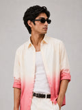 A man wearing a light cream and pink ombre button-down shirt over a white t-shirt, styled with white pants, a black belt, and black sunglasses, set against a plain light gray background.
