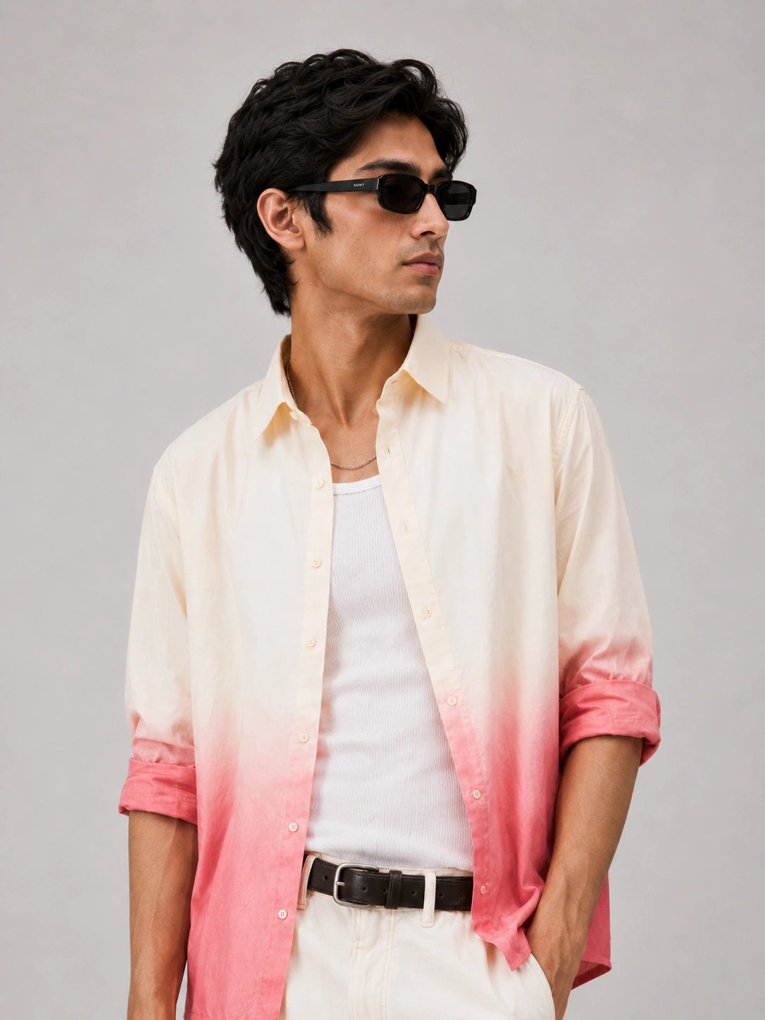 A man wearing a light cream and pink ombre button-down shirt over a white t-shirt, styled with white pants, a black belt, and black sunglasses, set against a plain light gray background.