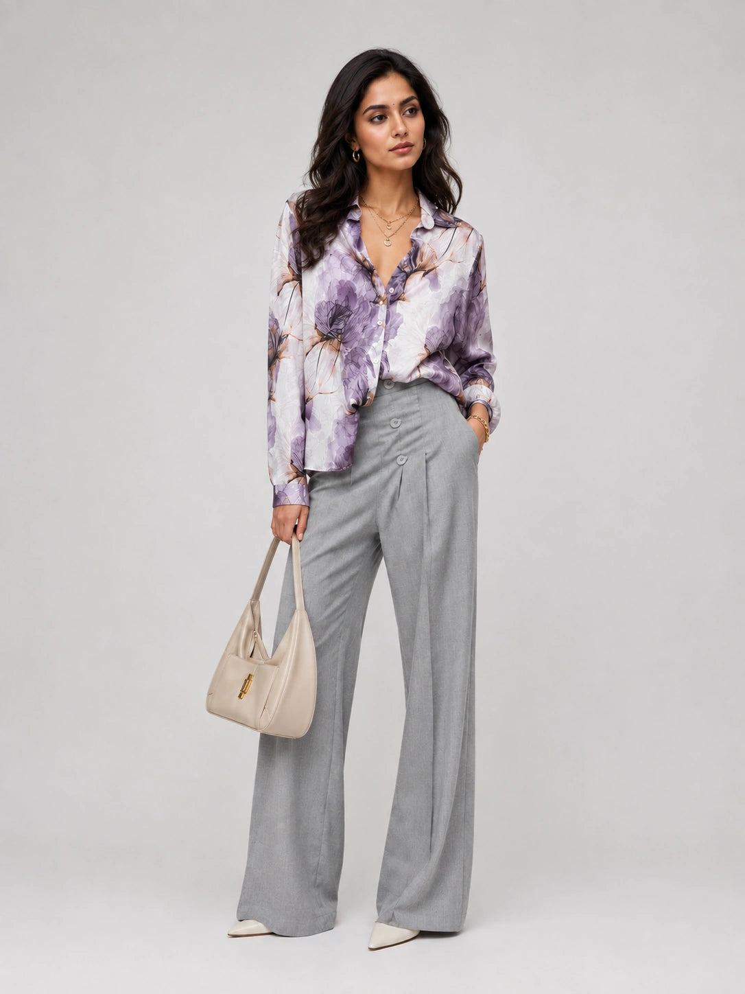 A woman models a long-sleeve button-up shirt with a purple, white, and brown watercolor floral pattern, paired with high-waisted light gray wide-leg trousers and a beige handbag, set against a plain light gray studio background.
