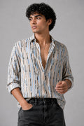 A man wearing a long-sleeve button-up shirt with a vertical stripe pattern in beige, blue, and black, paired with dark gray jeans, photographed against a solid gray studio background.