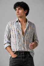 A man wearing a long-sleeve button-up shirt with a vertical stripe pattern in beige, blue, and black, paired with dark gray jeans, photographed against a solid gray studio background.