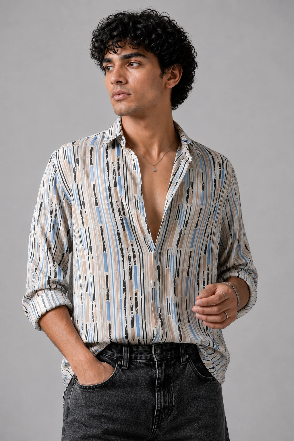 A man wearing a long-sleeve button-up shirt with a vertical stripe pattern in beige, blue, and black, paired with dark gray jeans, photographed against a solid gray studio background.